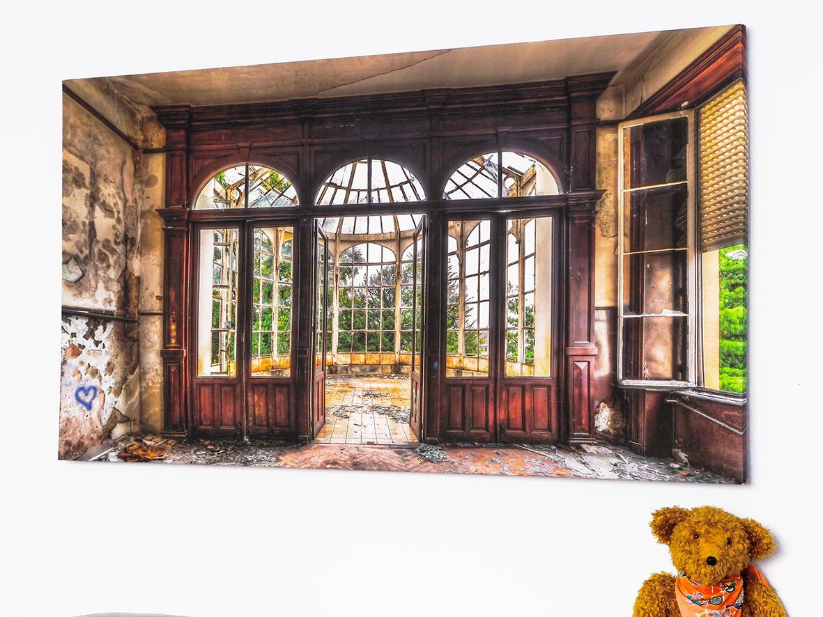 Verlassenes Zimmer mit großen Bogenfenstern, abblätternden Wänden und einem Teddybären auf dem Boden.