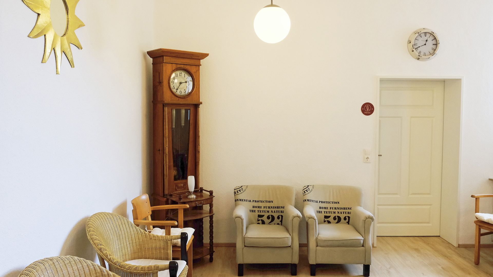 Helles Wohnzimmer mit Sesseln, Uhrenschrank, Hängelampen und einer weißen Tür auf der rechten Seite.