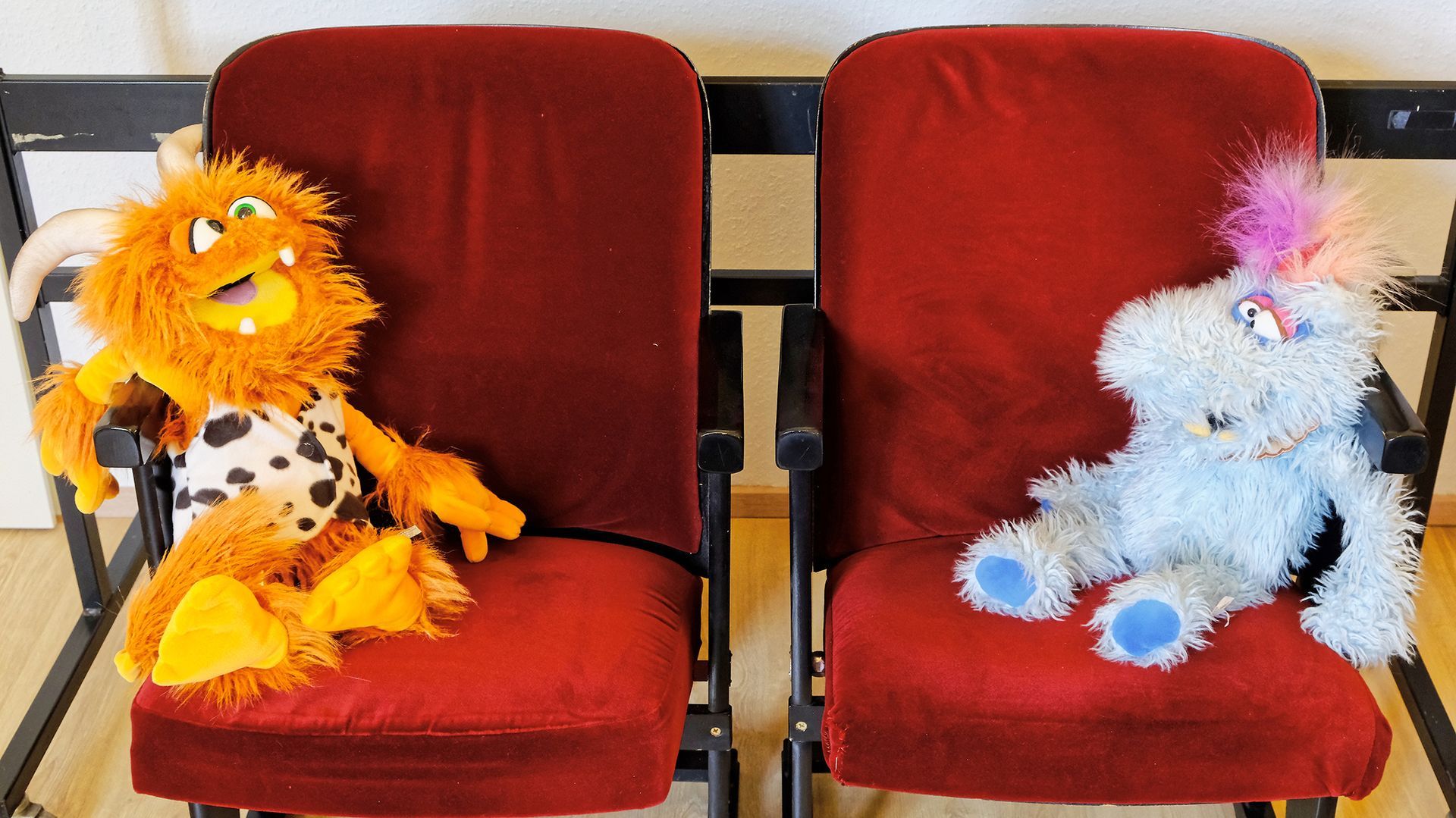 Ein orangefarbener Plüschlöwe und ein blauer Plüschhase sitzen auf roten Stühlen vor einer gestreiften Wand.