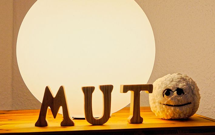 Holzbuchstaben, die „MUT“ bilden, neben einem kleinen, flauschigen Gesicht unter einer leuchtenden runden Lampe