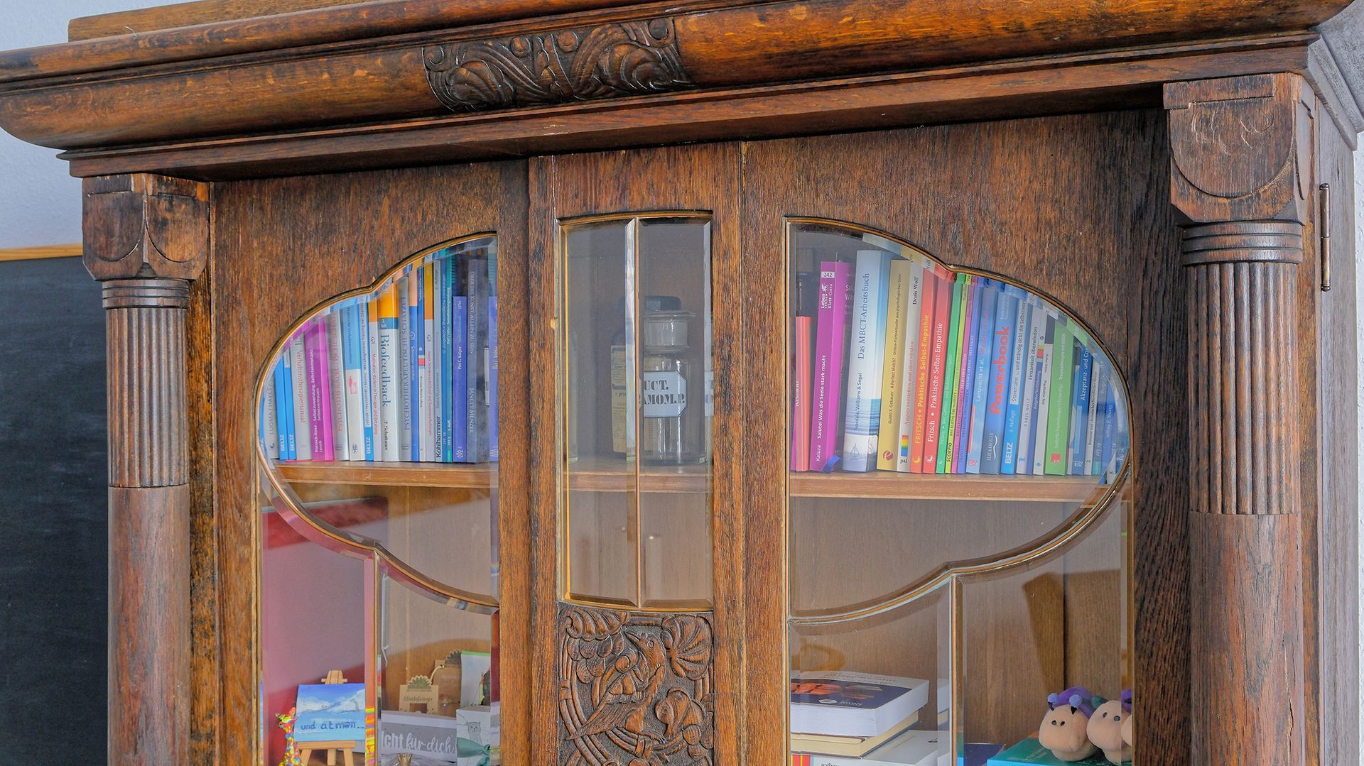 Ein reich verzierter Holzschrank mit Glastüren, in dem sich farbenfrohe Bücher und Keramiken befinden.