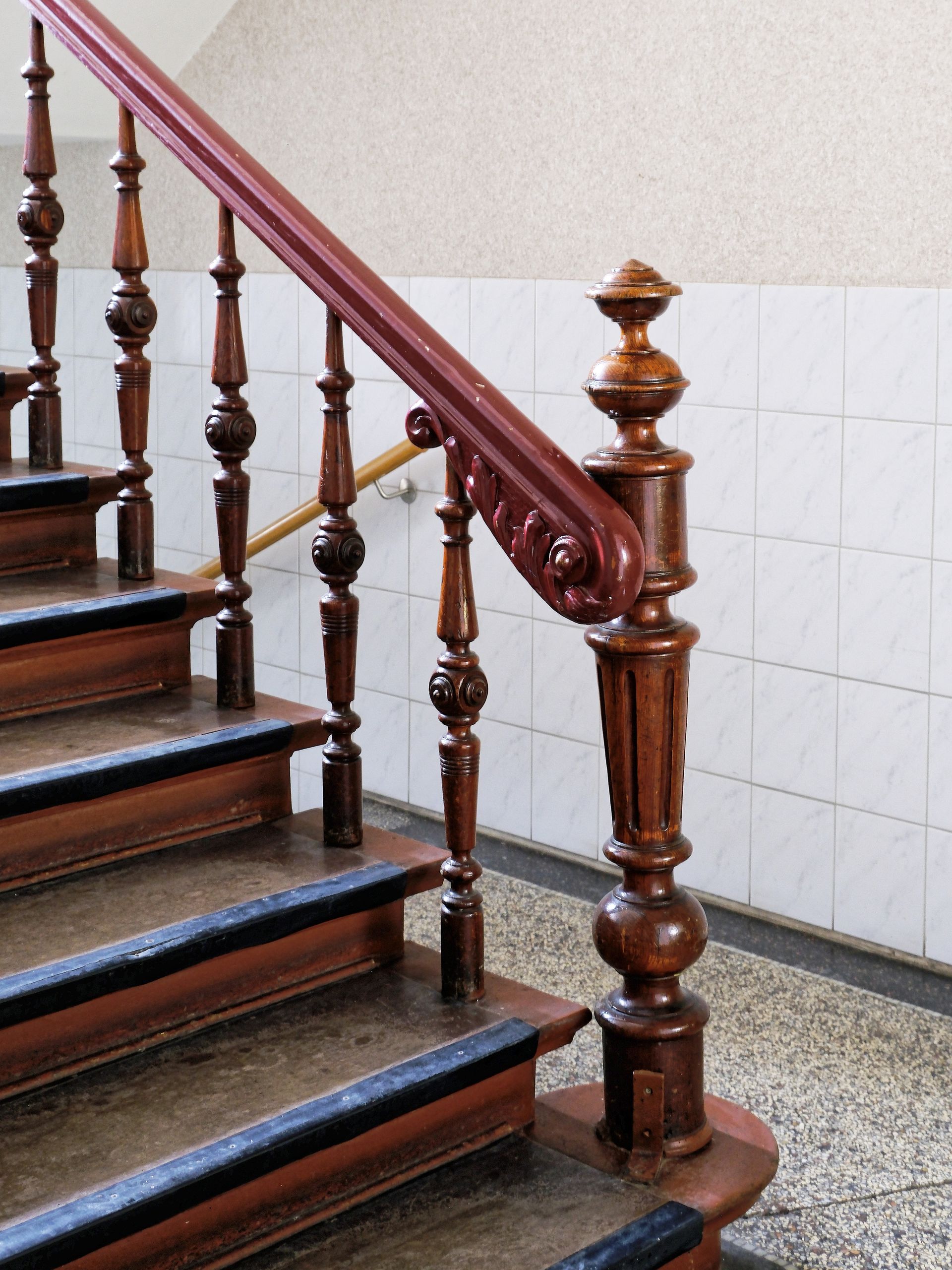 Holztreppe mit dunkelrotem Handlauf und geschnitztem Treppengeländer neben einer gefliesten Wand