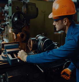 Ouvrier en combinaison bleue manipulant des machines, portant des lunettes de sécurité et un casque orange, dans un environnement industriel.