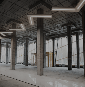Intérieur en béton en cours de construction avec piliers de soutien, grandes fenêtres et luminaires géométriques au plafond.