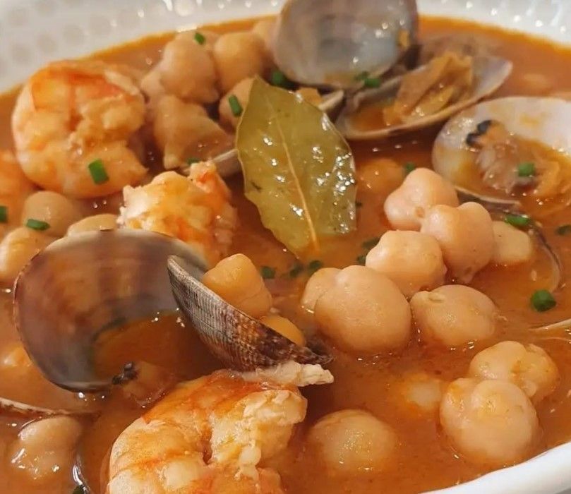 Guiso de mariscos con camarones, almejas, garbanzos y laurel en un recipiente blanco.