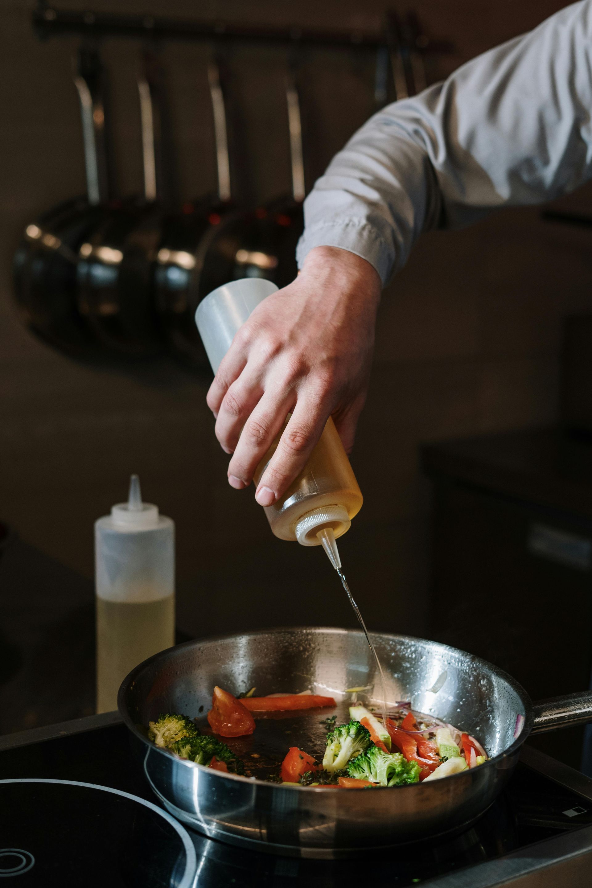 Chef vertiendo aceite en una sartén con verduras sobre una estufa en una cocina.