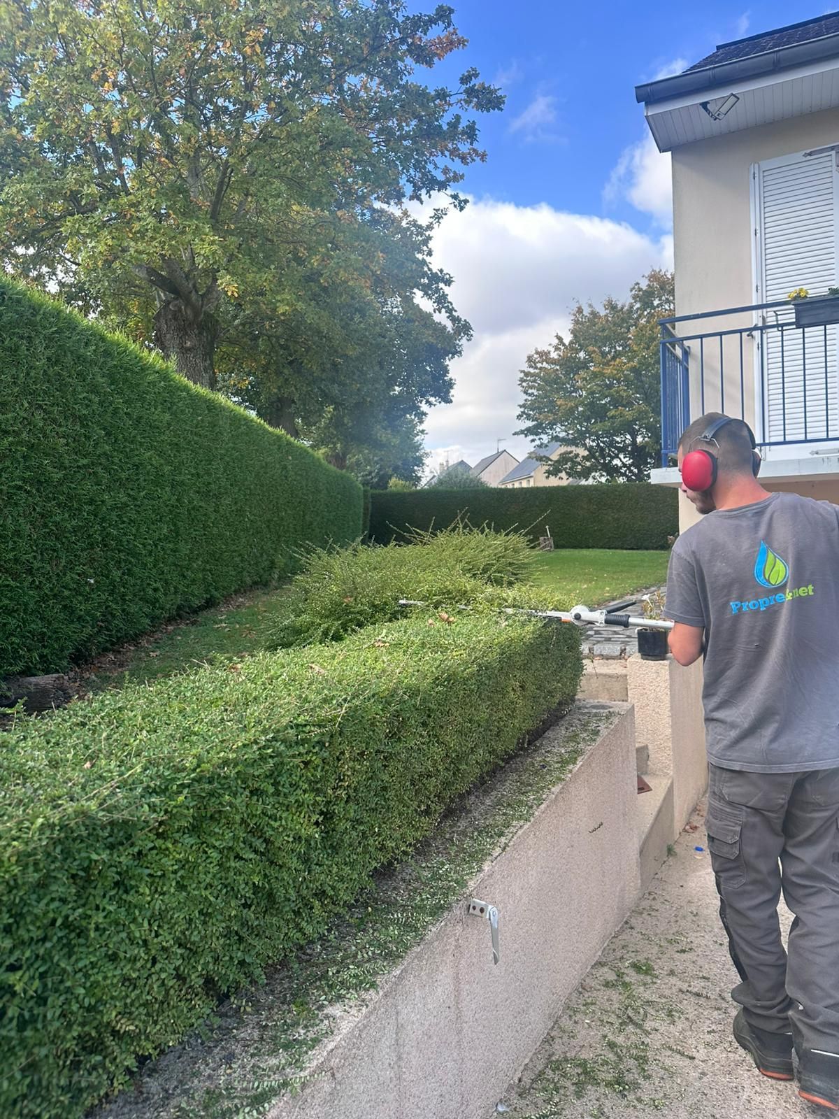 Un homme taille des haies avec une scie électrique près d'un bâtiment et d'autres espaces verts sous un ciel nuageux.