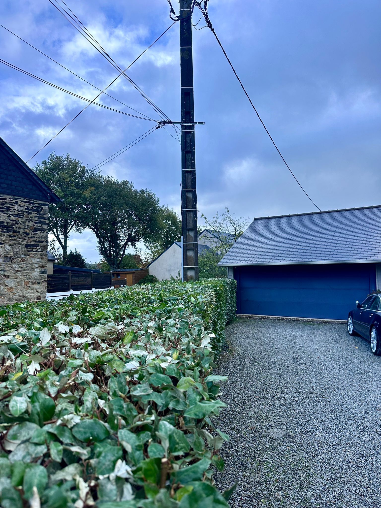 Lignes électriques au-dessus d'une allée de gravier, d'un garage et d'une haie, par temps nuageux.