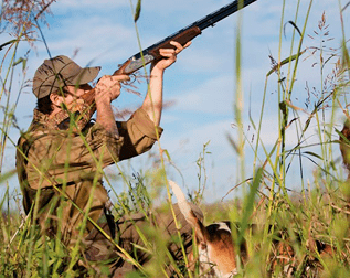 Cazador apuntando con una escopeta hacia arriba en la hierba alta bajo un cielo azul