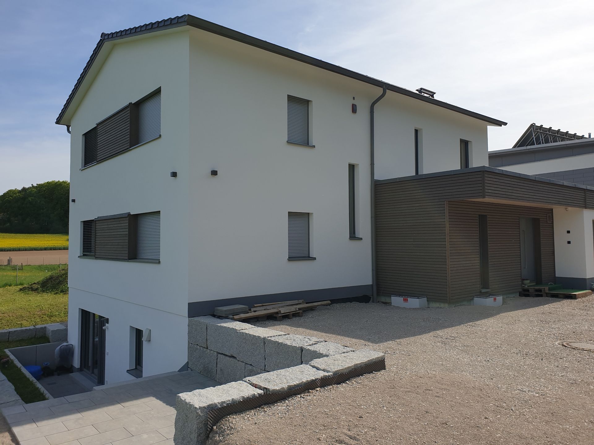 Weißes zweistöckiges Haus mit schwarzem Dach, grauer Stützmauer und Kiesauffahrt.