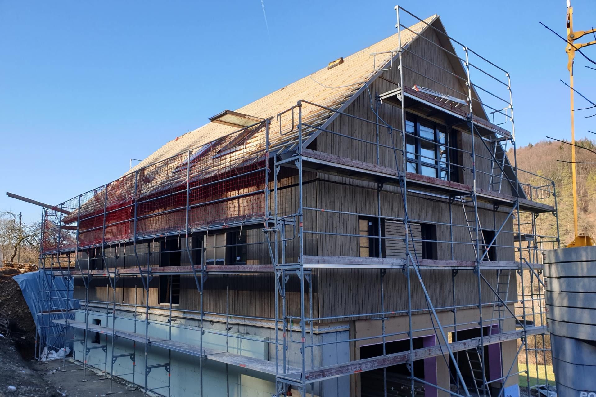 Im Bau befindliches Holzhaus, umgeben von Gerüsten, an einem sonnigen Tag.