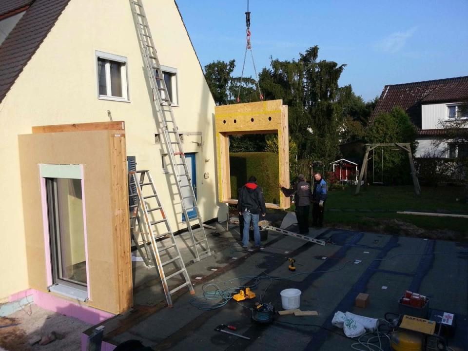 Bauarbeiter installieren mit einem Kran einen Holzrahmenanbau an einem beigen Haus.
