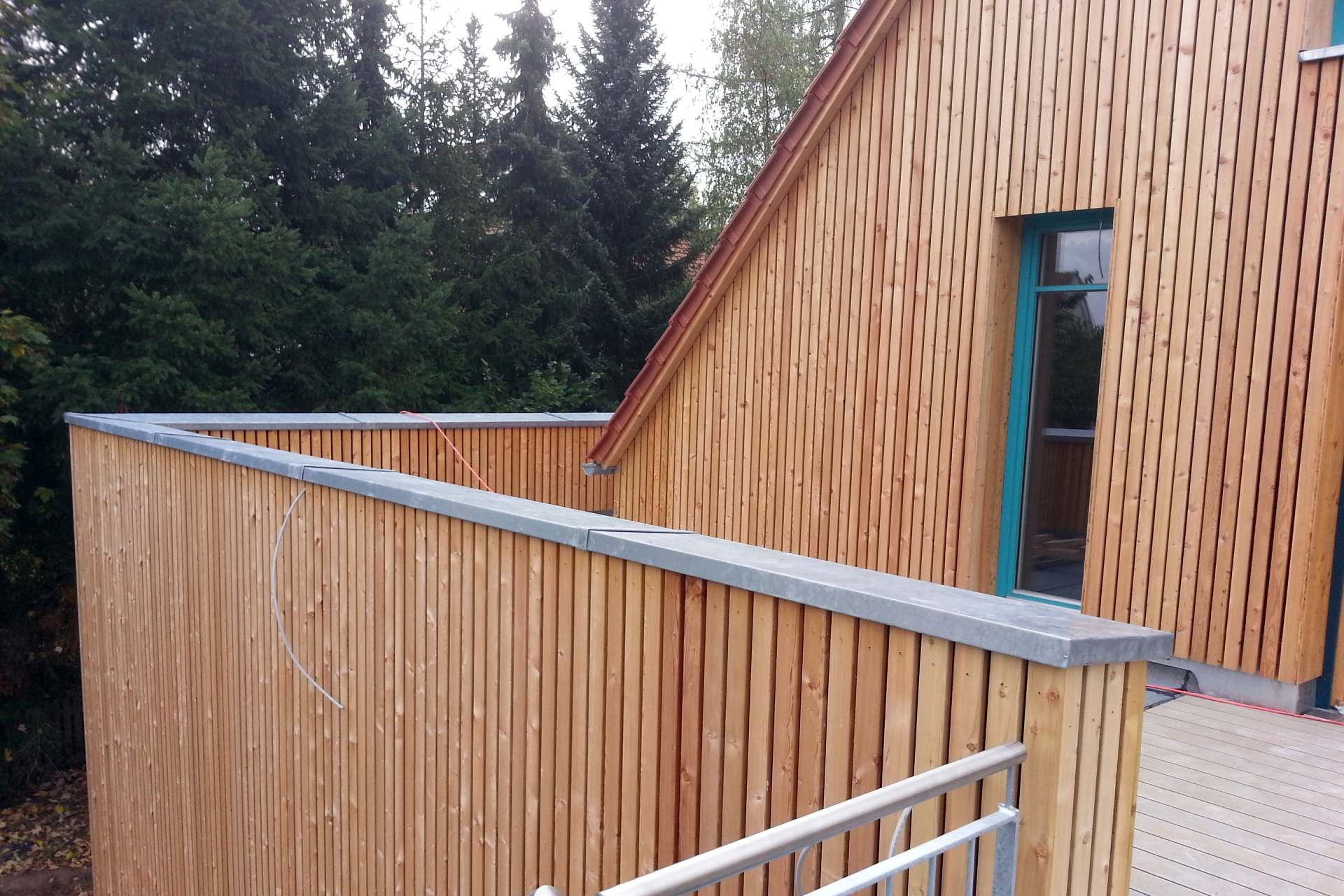 Holzgebäude mit Balkon und gemusterter Außenverkleidung neben grünen Bäumen.