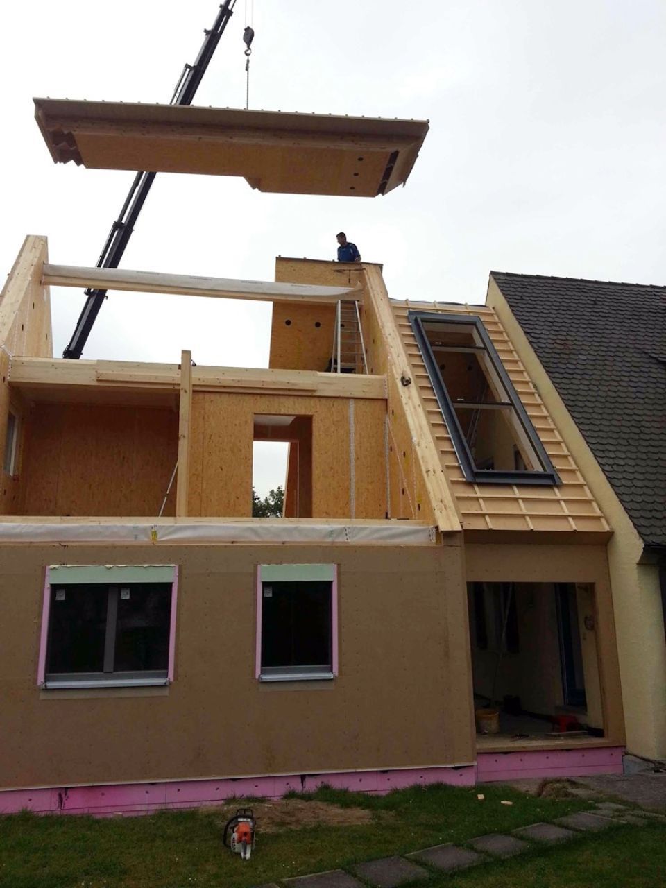 Ein Kran hebt ein hölzernes Dachelement auf ein im Bau befindliches Haus; ein Arbeiter steht auf dem Dach.
