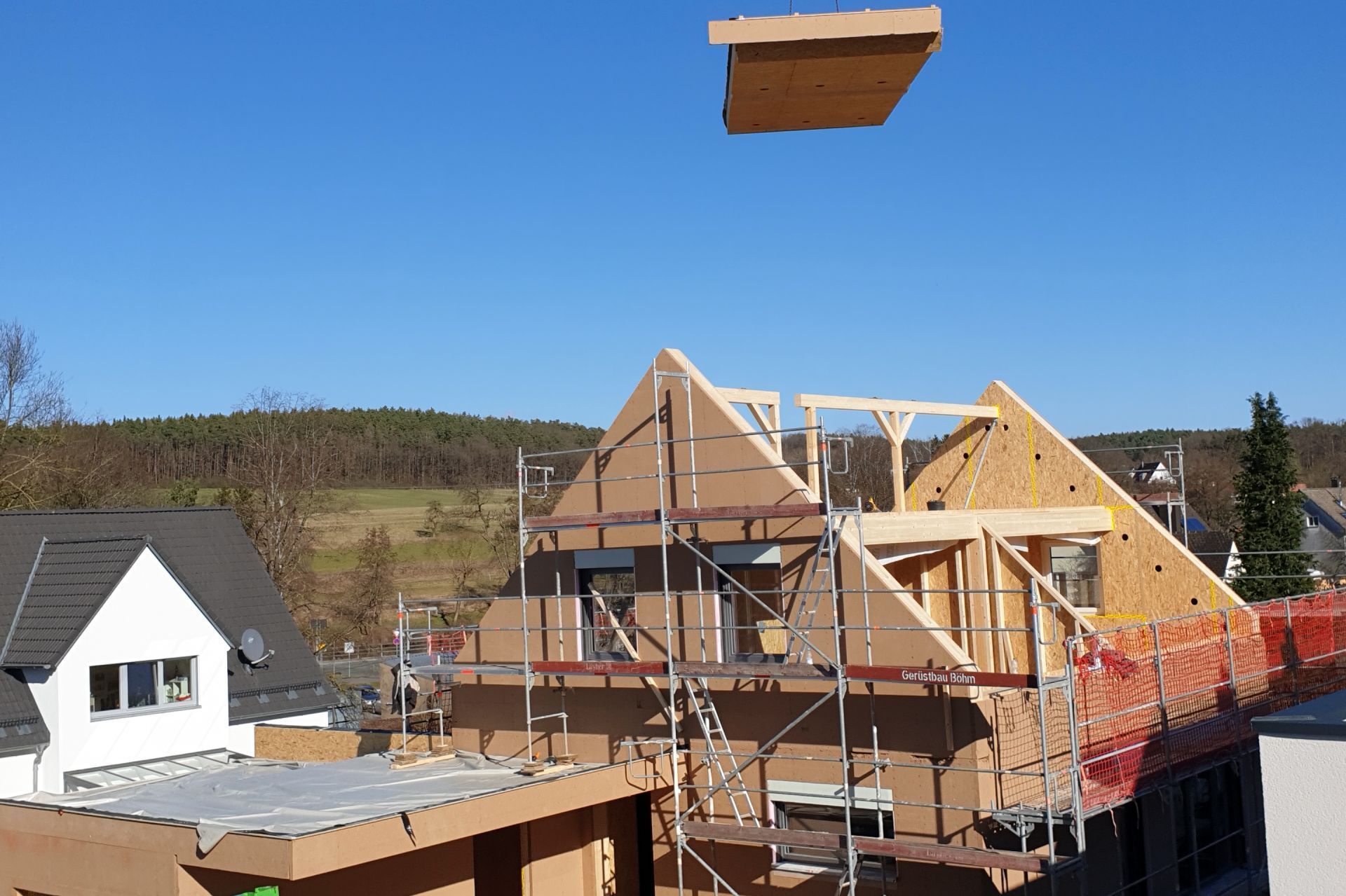 Baustelle mit Hausrahmen, Gerüst und Holzplatte, die vor einem blauen Himmel angehoben wird.