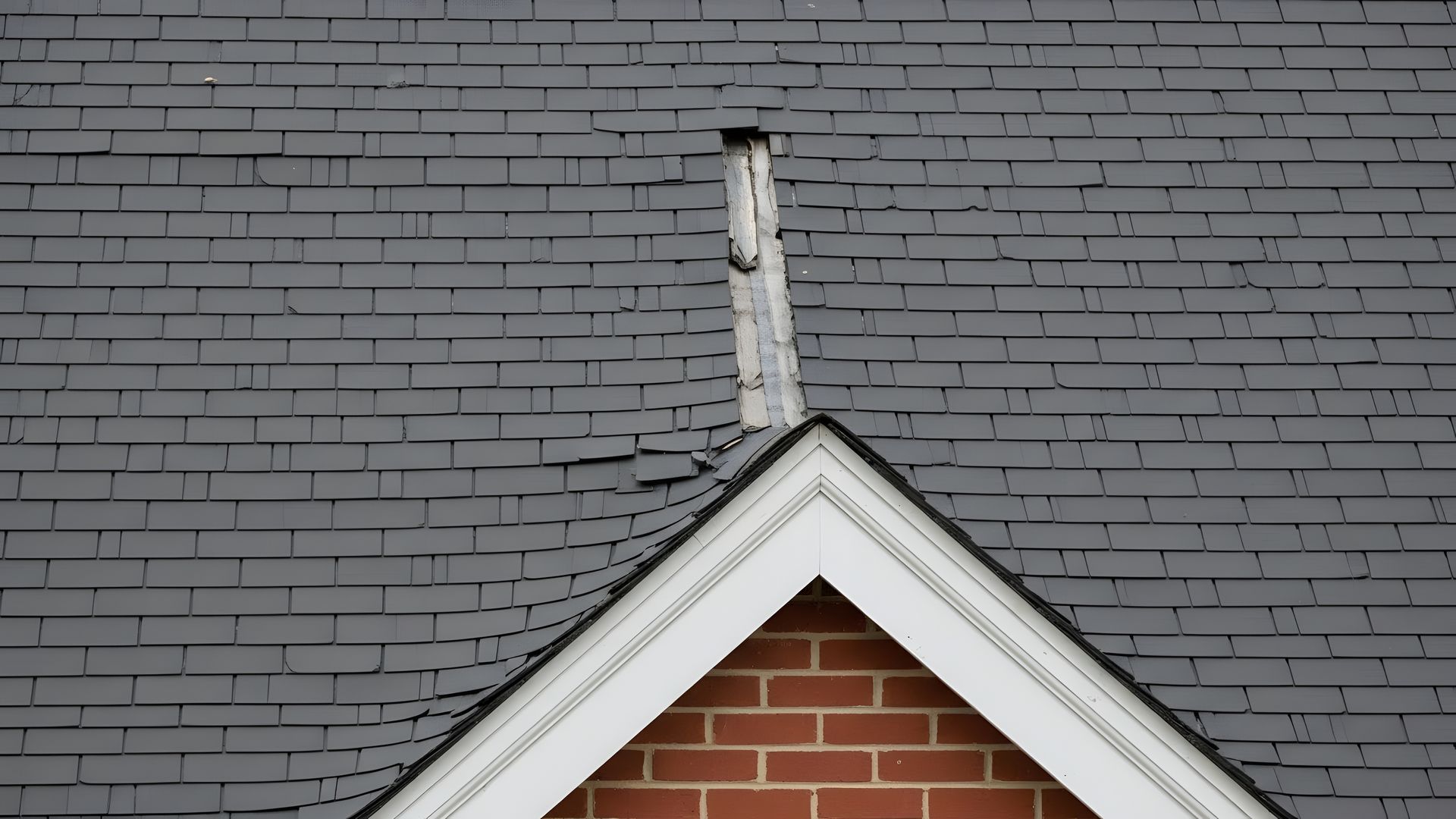 Gros plan sur un toit en bardeaux gris avec un triangle bordé de blanc et un mur de briques en dessous.