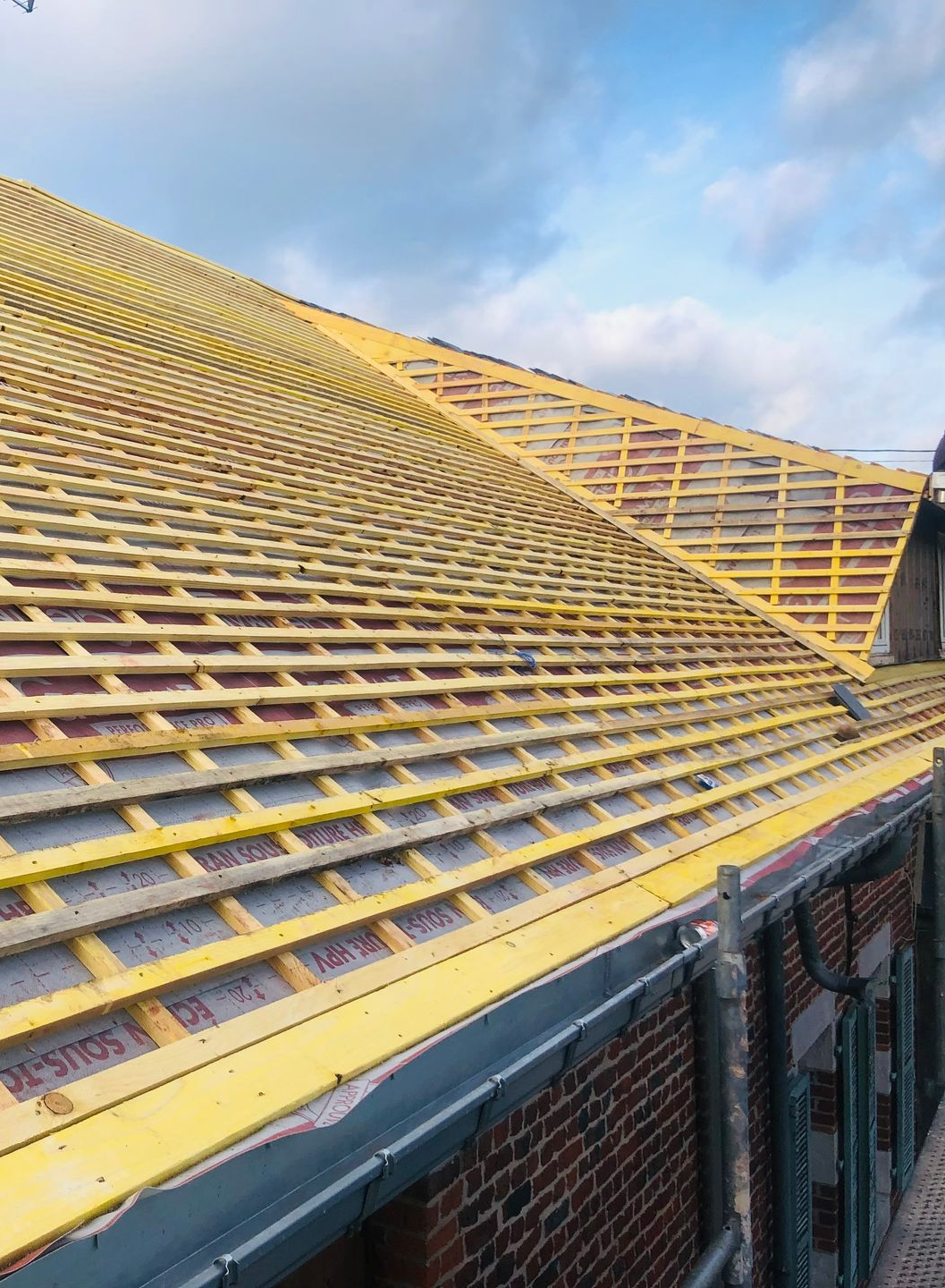 Vue d'une toiture en cours de construction, avec des chevrons en bois, des poutres jaunes et un mur de briques.