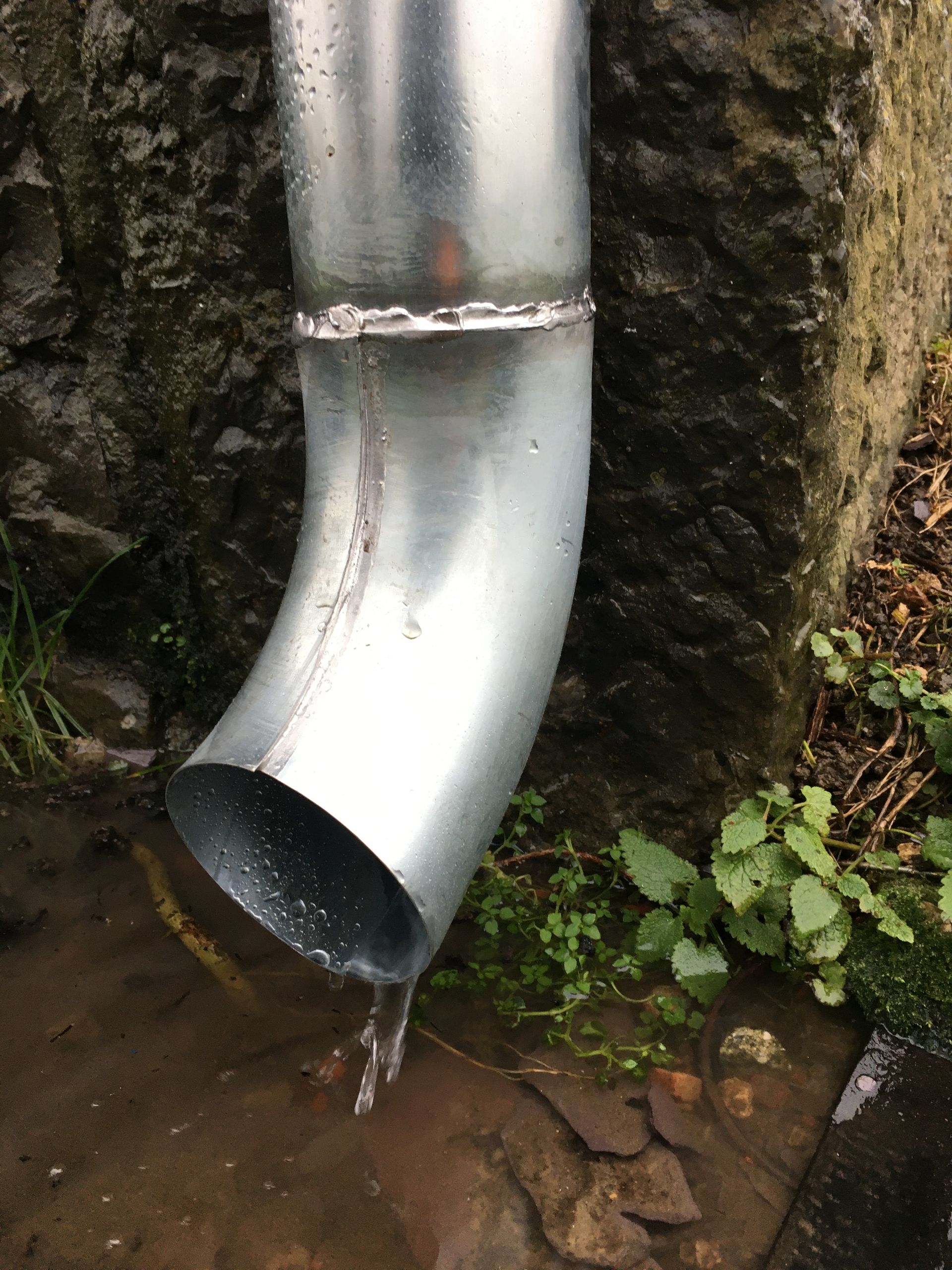 Une descente de gouttière métallique fuit contre un mur en pierre.