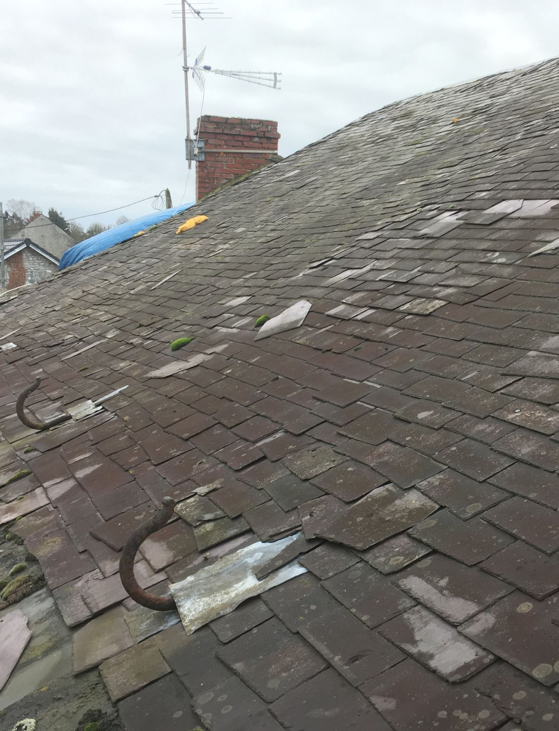 Un vieux toit d'ardoise avec des crochets métalliques, une cheminée et une antenne au loin.