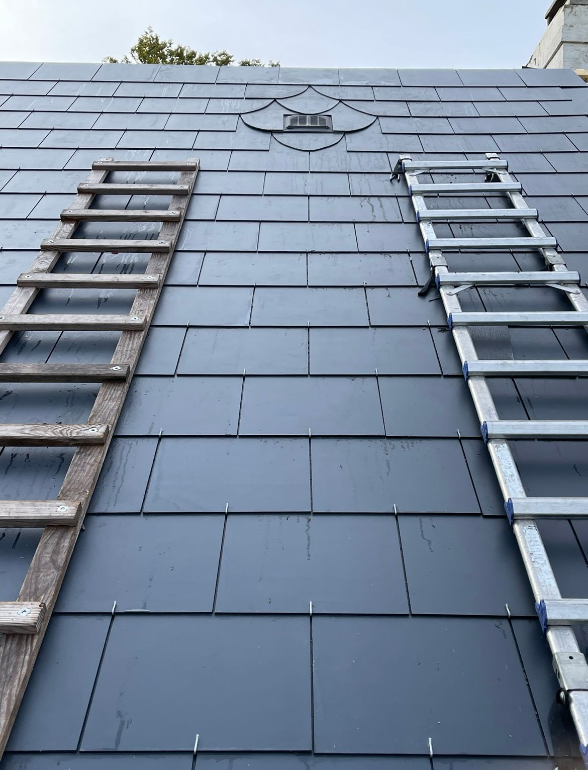 Deux échelles appuyées contre un toit d'ardoise gris foncé. Le toit est orné d'un élément décoratif en forme de losange.