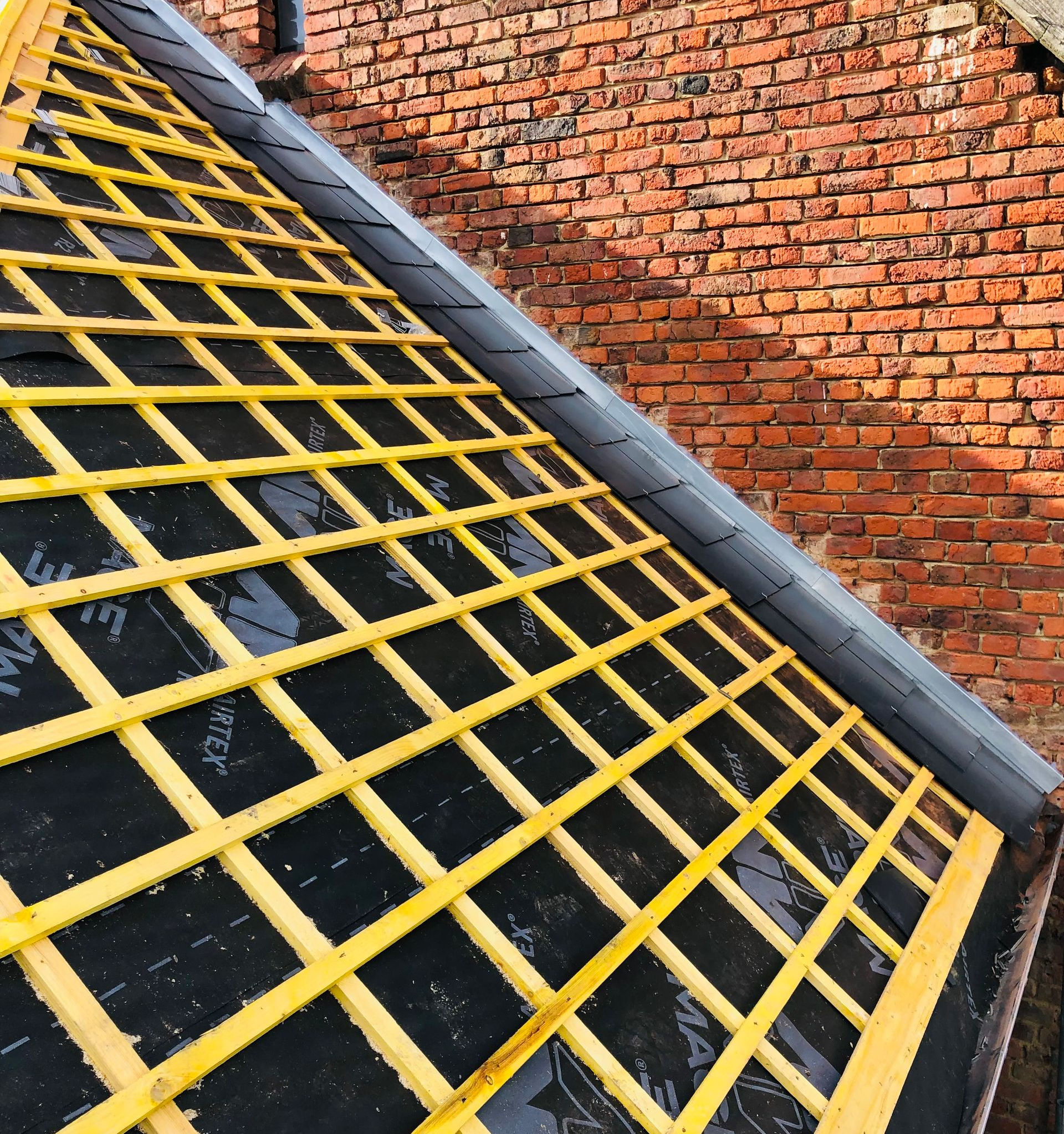 Charpente de toit en bois avec sous-couche noire, à côté d'un mur en briques rouges.