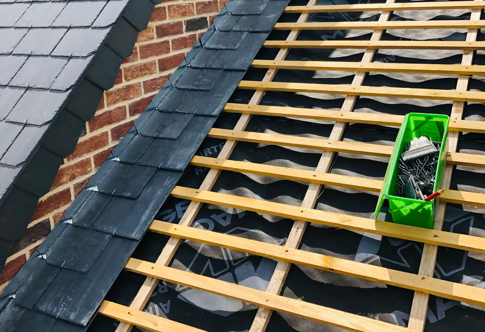 Toiture en construction : bardeaux d’ardoise foncée, charpente en bois, section de mur en briques et outils dans un bac vert.