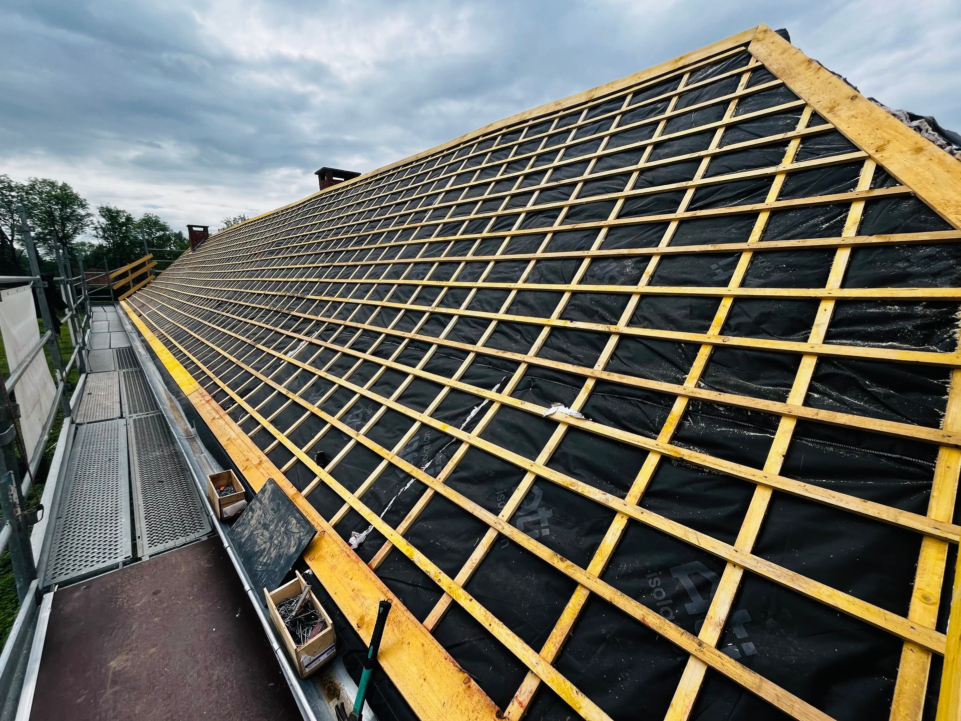 Toiture en construction, sous-couche noire, supports en bois, échafaudages sur le côté, ciel couvert.