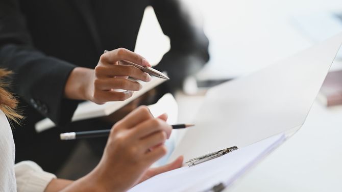 Zwei Personen in Geschäftskleidung prüfen Dokumente, eine von ihnen zeigt mit einem Stift darauf.
