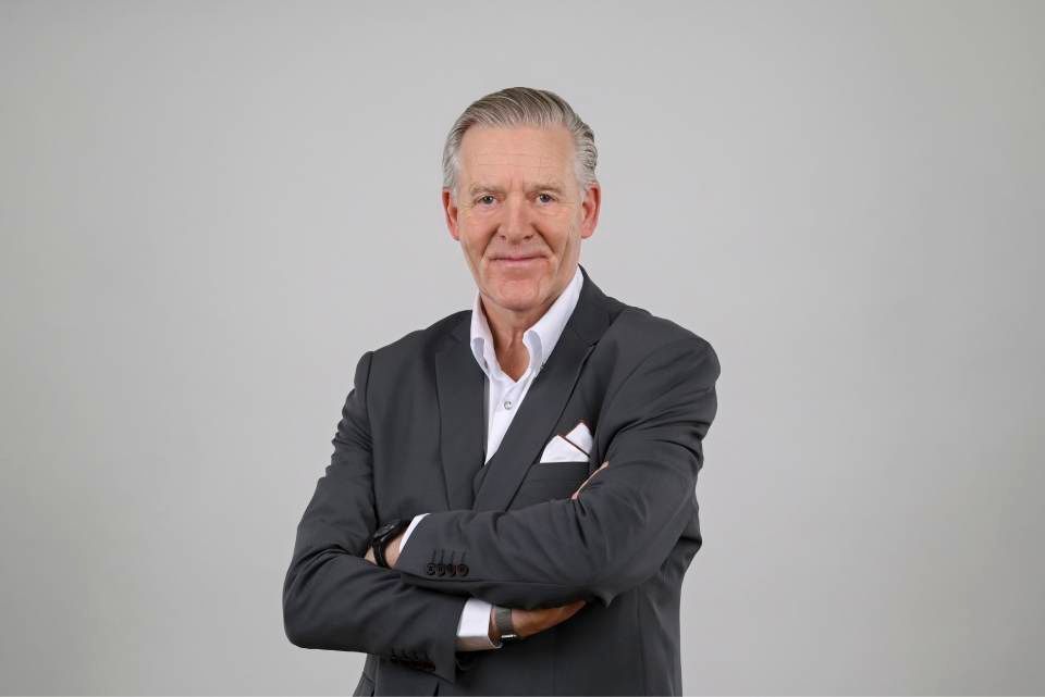 Mann im dunklen Anzug mit verschränkten Armen, lächelnd, vor grauem Hintergrund.