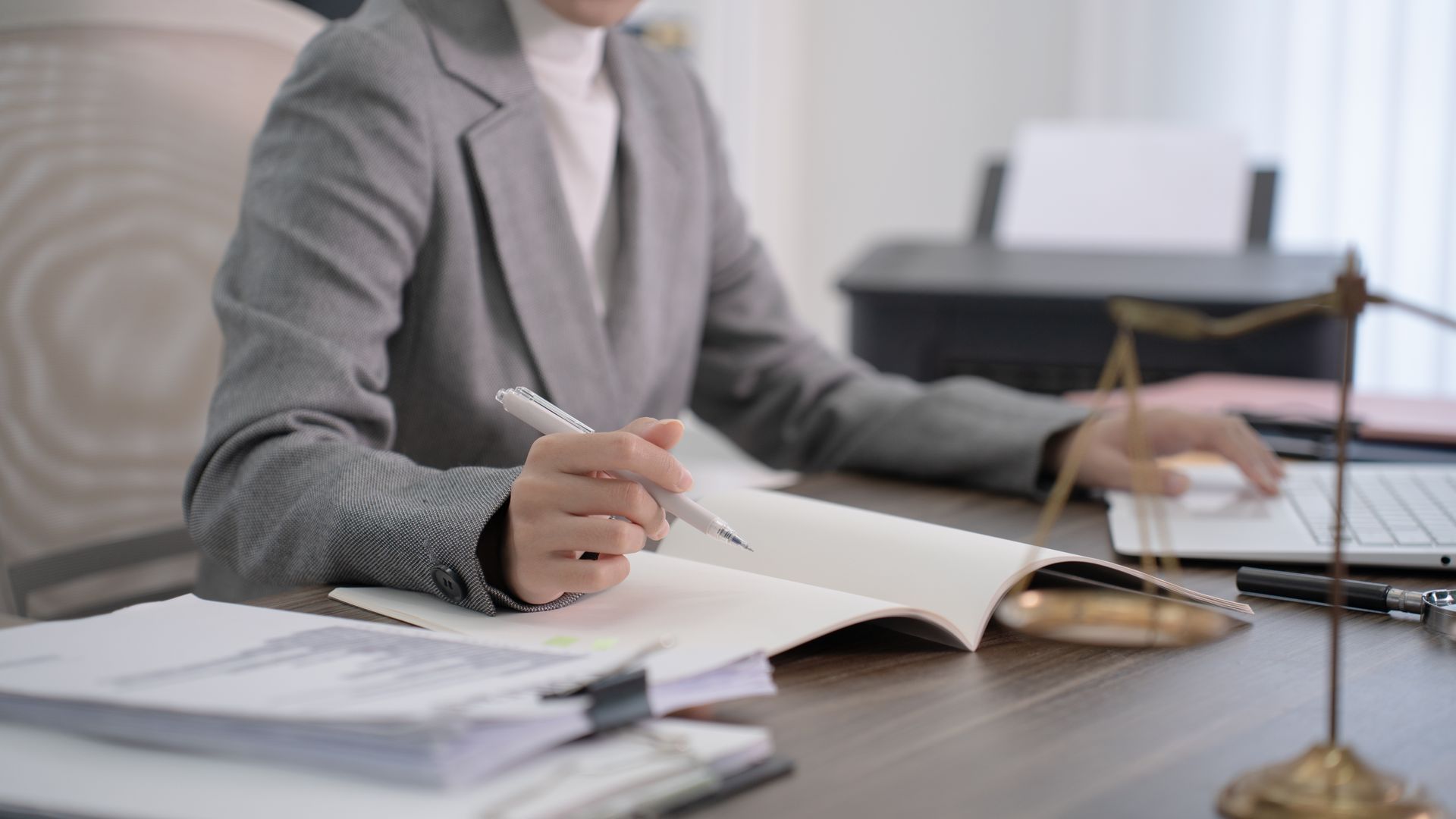 Une personne en blazer gris signe des documents à un bureau où s'empilent des papiers et où se trouve une balance.