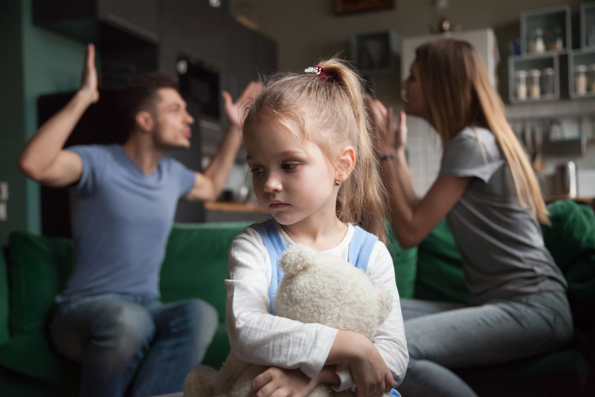 Un jeune enfant tenant un ours en peluche, tandis que des adultes se disputent en arrière-plan.