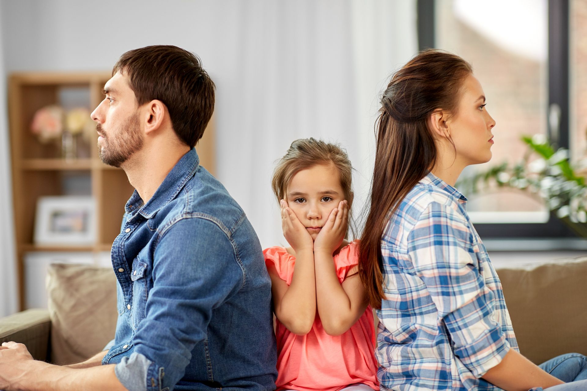 Une famille assise à distance les unes des autres sur un canapé, un enfant au milieu, les mains sur les joues, dans un salon.