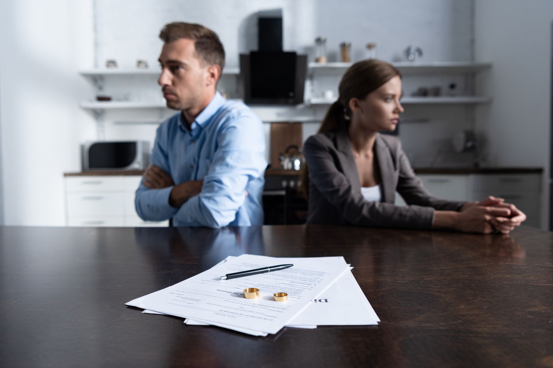 Deux personnes sont assises dos à dos à une table dans une cuisine, avec des papiers de divorce au premier plan.