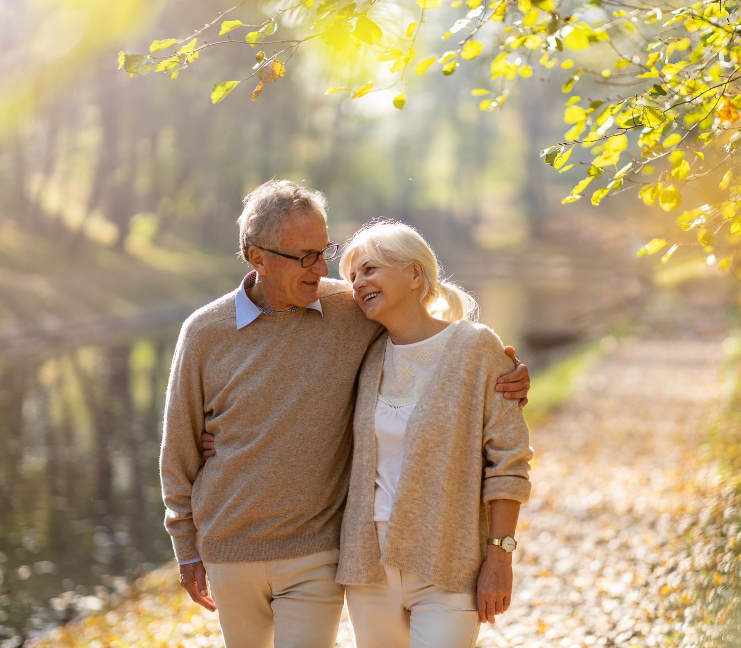 Un couple souriant se promenant bras dessus bras dessous sous des arbres baignés de soleil dans un parc.
