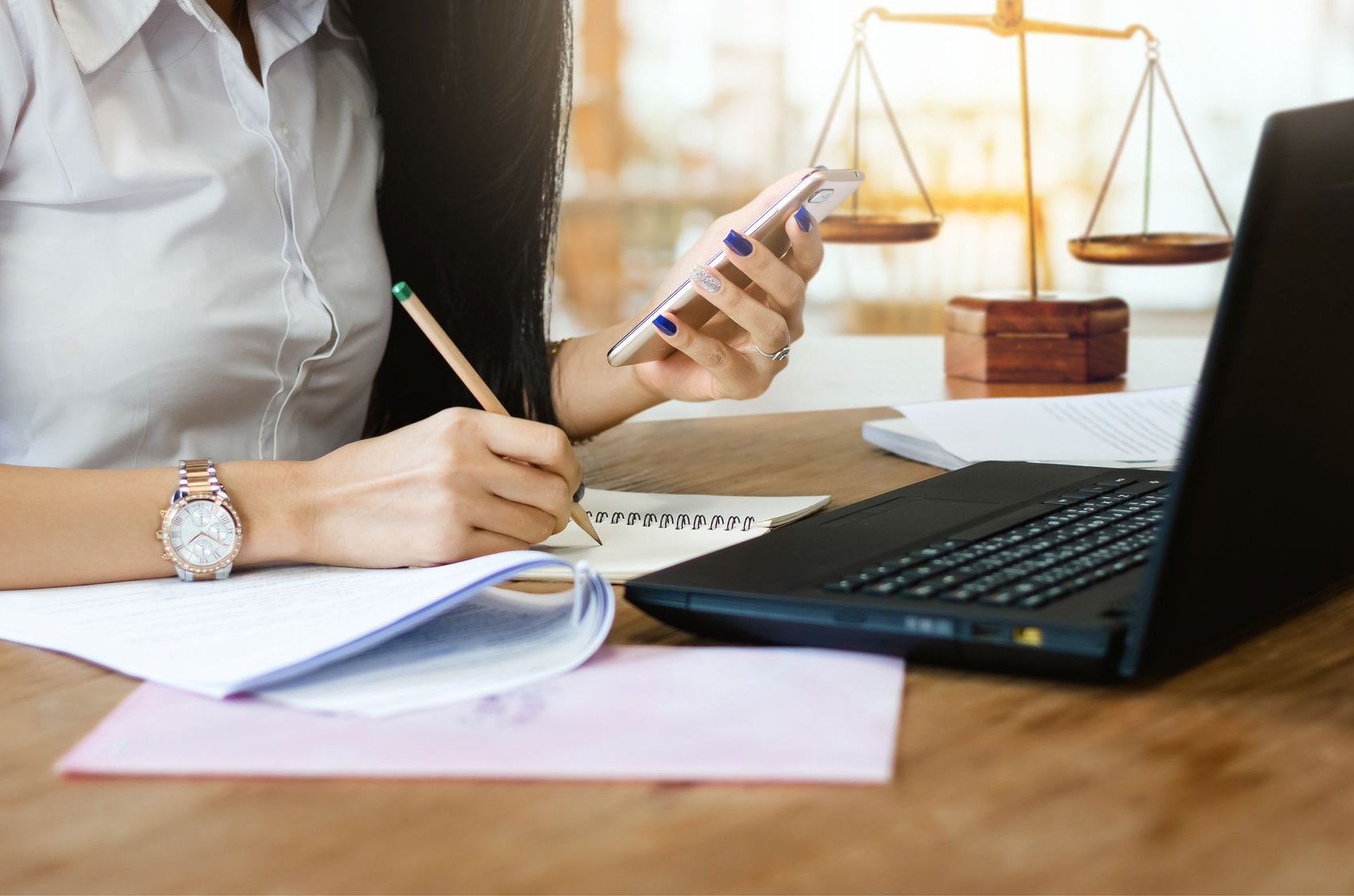 Une personne écrit dans un cahier à côté d'un ordinateur portable tout en tenant un smartphone sur son bureau.
