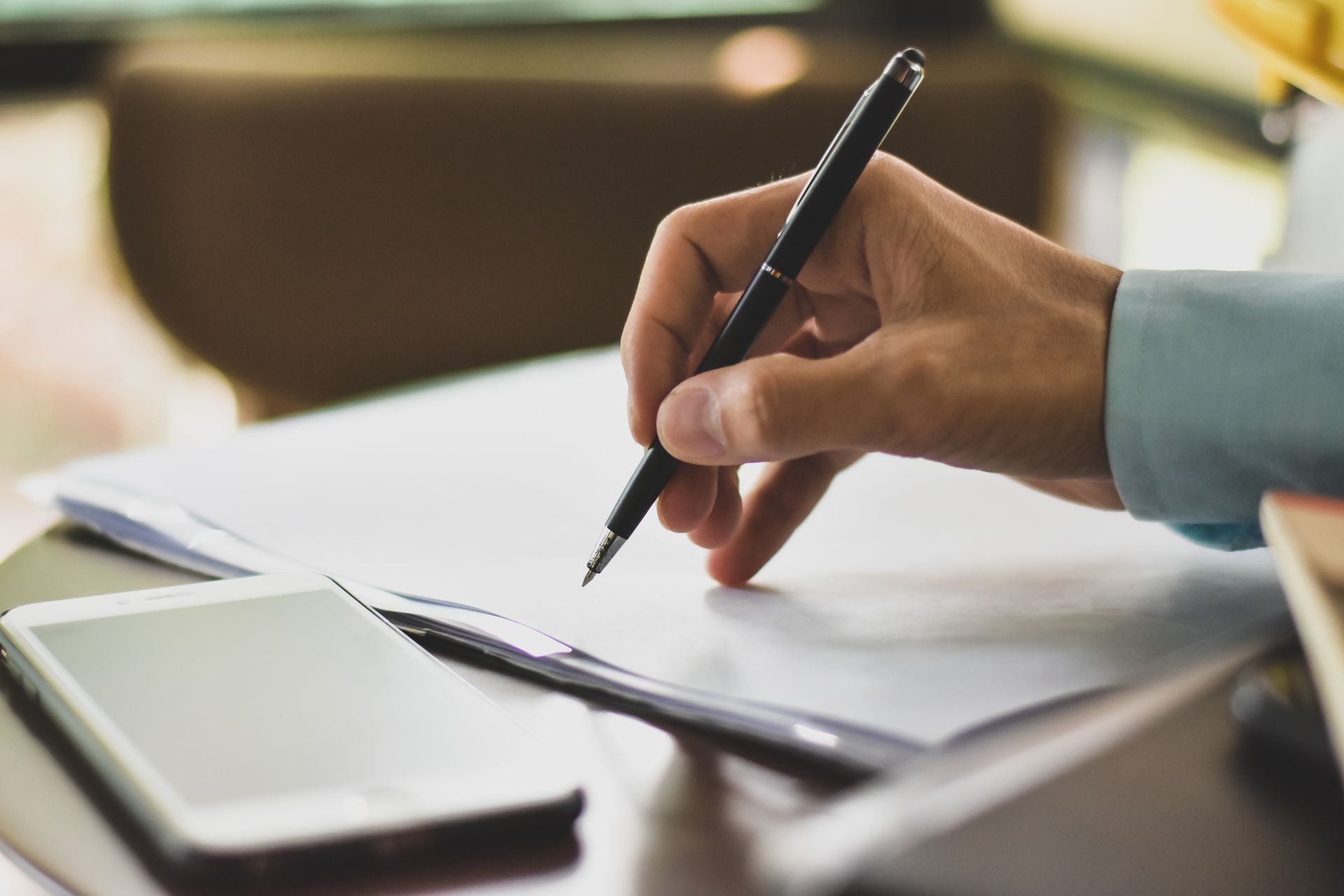 Une main tenant un stylo au-dessus d'un cahier à côté d'un smartphone posé sur un bureau.