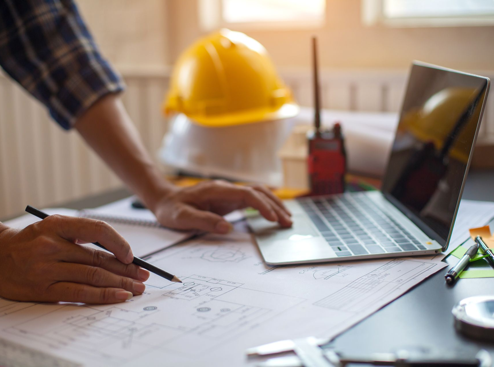 Une personne dessine des plans à un bureau avec un ordinateur portable, un casque de chantier jaune et des fournitures de bureau.