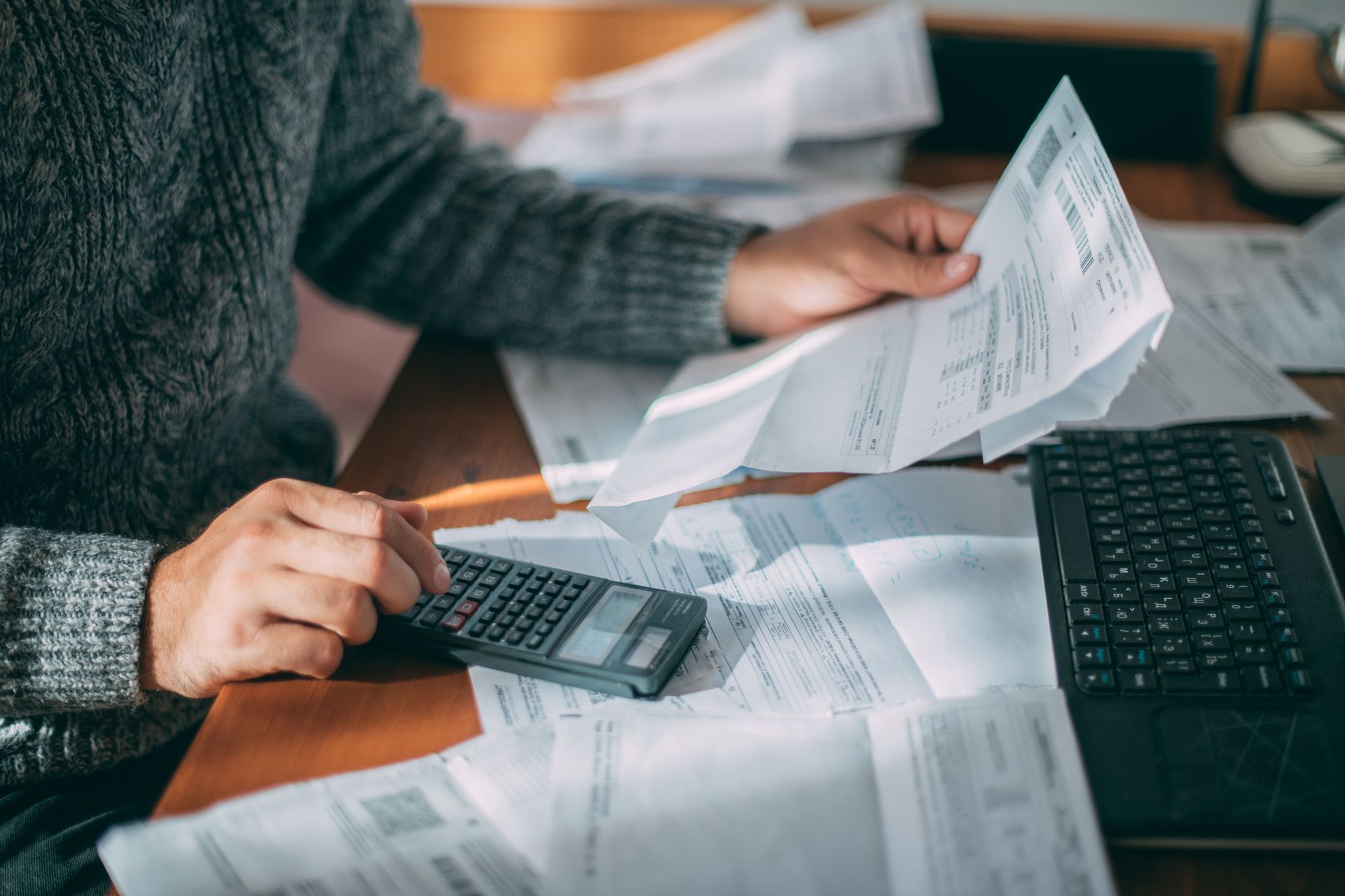 Des mains manipulant une calculatrice et triant des billets de banque à côté d'un ordinateur portable et de reçus éparpillés sur un bureau.
