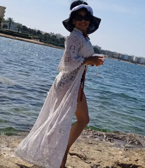 Una mujer con un pareo de encaje blanco y un sombrero para el sol está de pie cerca del agua sosteniendo una bebida.