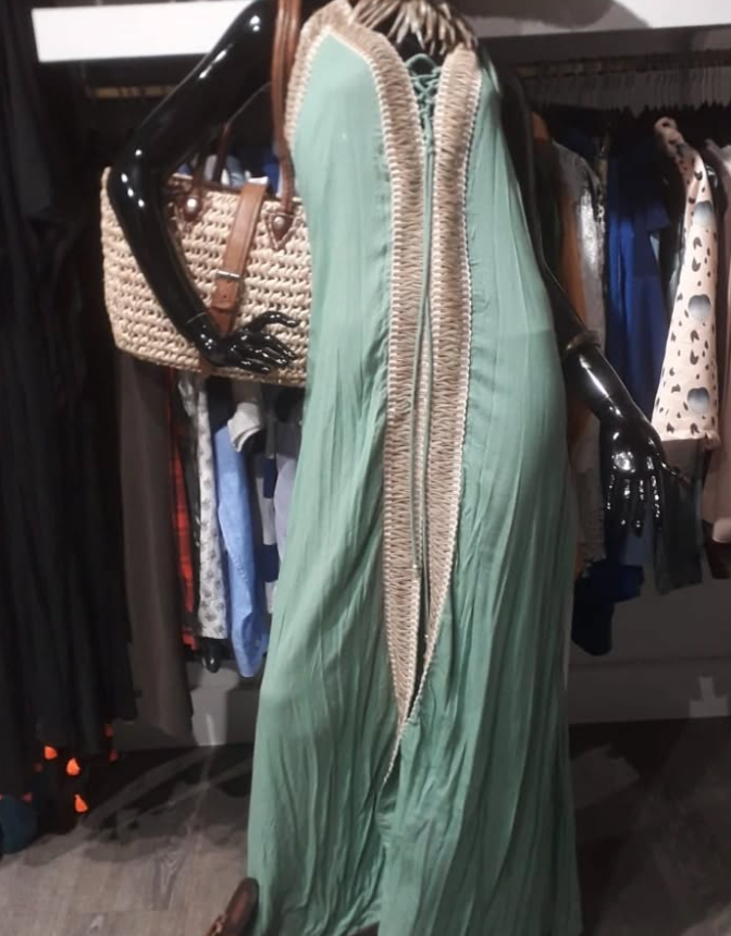 Un maniquí con un vestido verde menta con ribete beige, sosteniendo un bolso de paja; perchero al fondo.