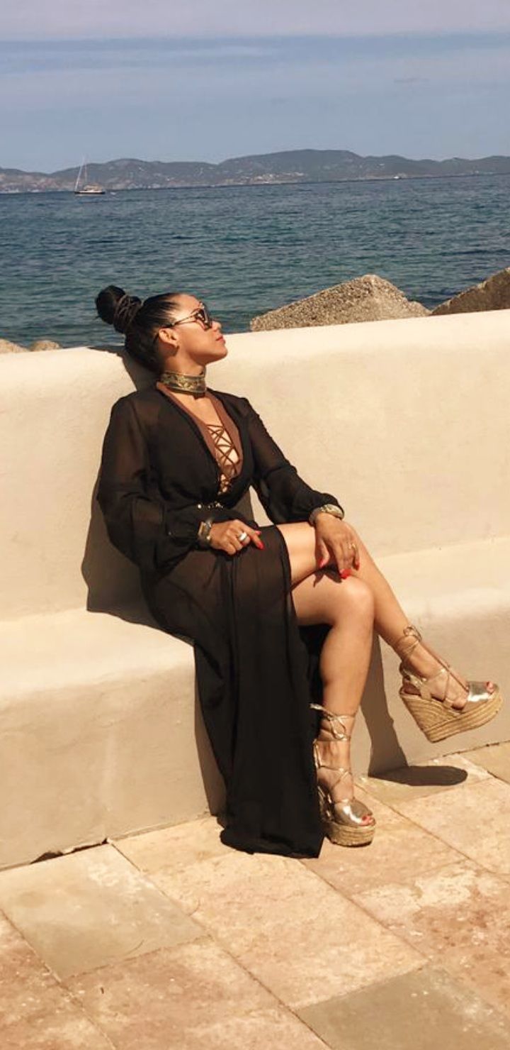 Mujer con vestido negro transparente y zapatos de plataforma, sentada junto al mar, mirando hacia arriba con gafas de sol.