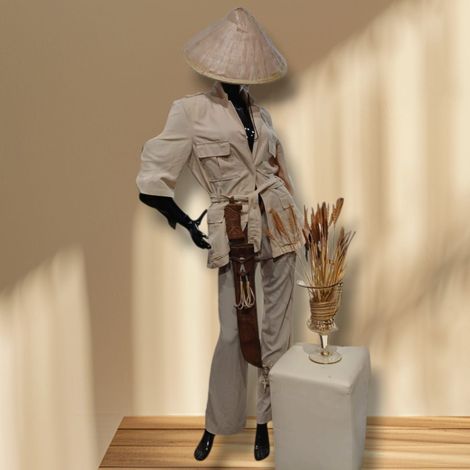 Maniquí con atuendo beige estilo safari, sombrero cónico y vestido ajustado, de pie junto a un jarrón con plantas secas.