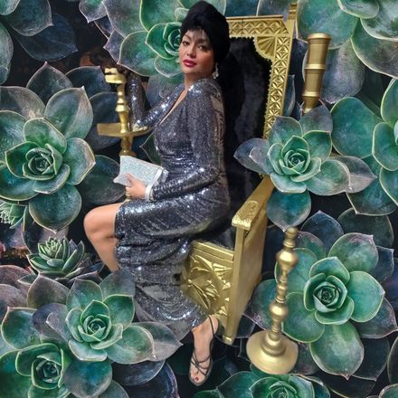Mujer con vestido de lentejuelas, sentada en un trono dorado, rodeada de plantas suculentas. Lleva un bolso de mano plateado.