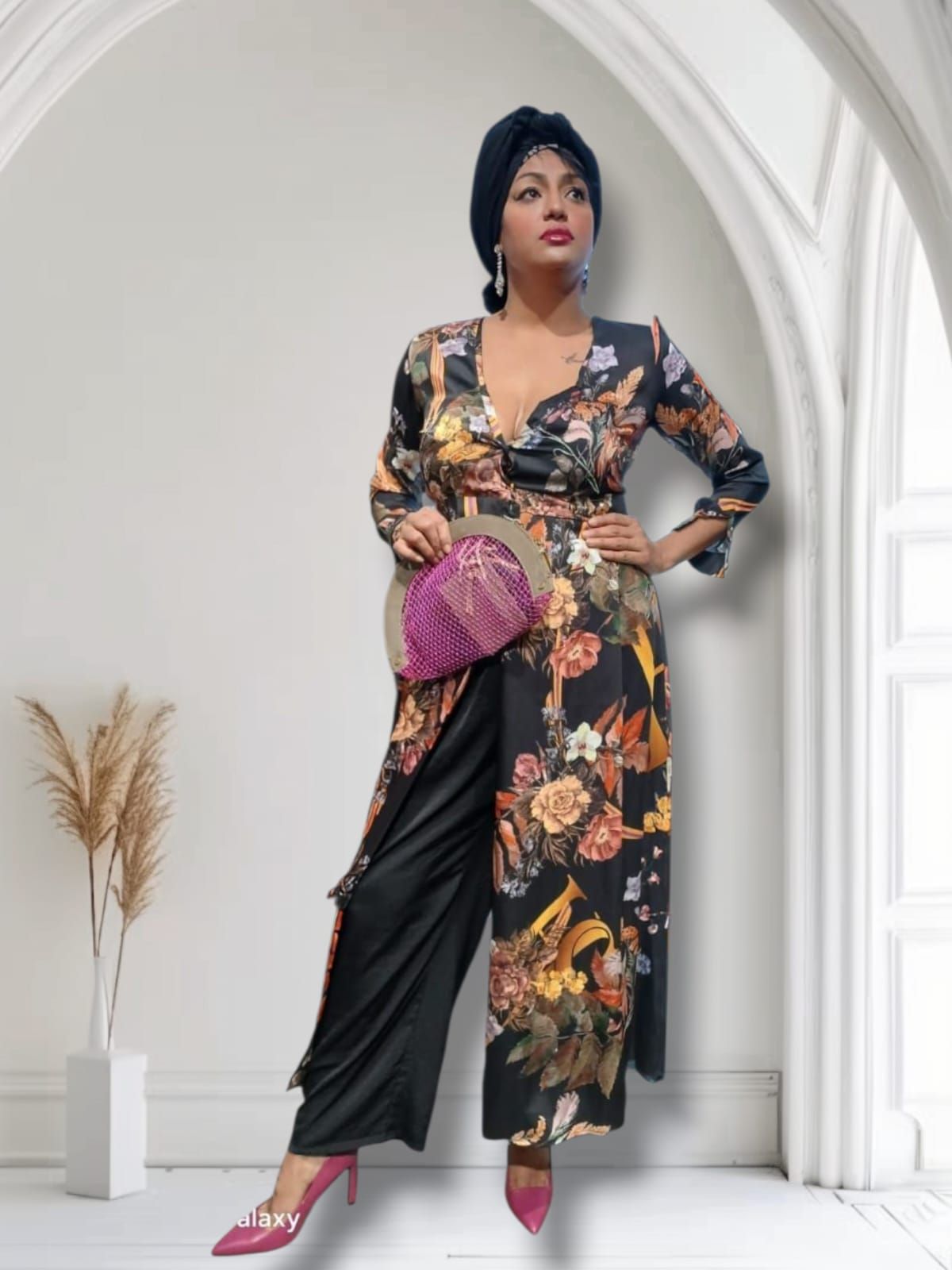 Mujer con atuendo floral y pañuelo en la cabeza, sosteniendo un bolso de mano rosa, posando frente a una ventana arqueada.