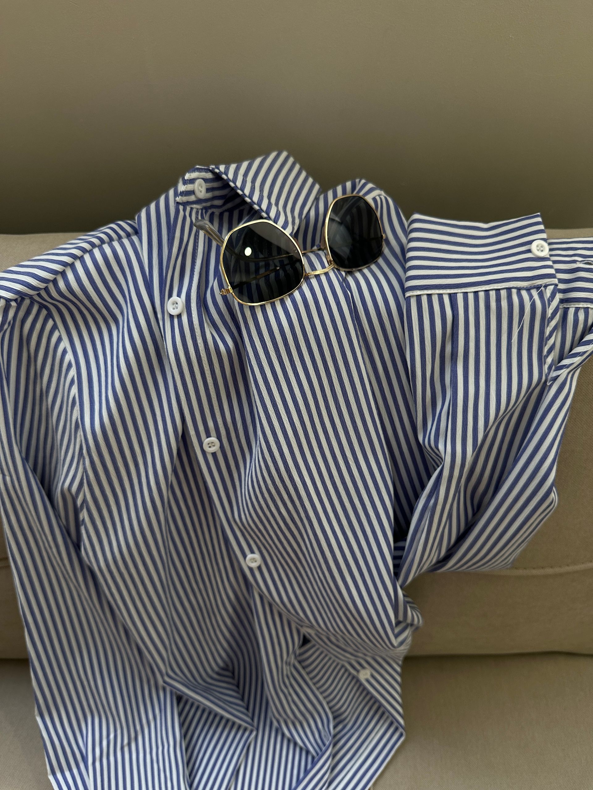 Camisa abotonada de rayas azules y blancas con gafas de sol sobre una superficie de color neutro.