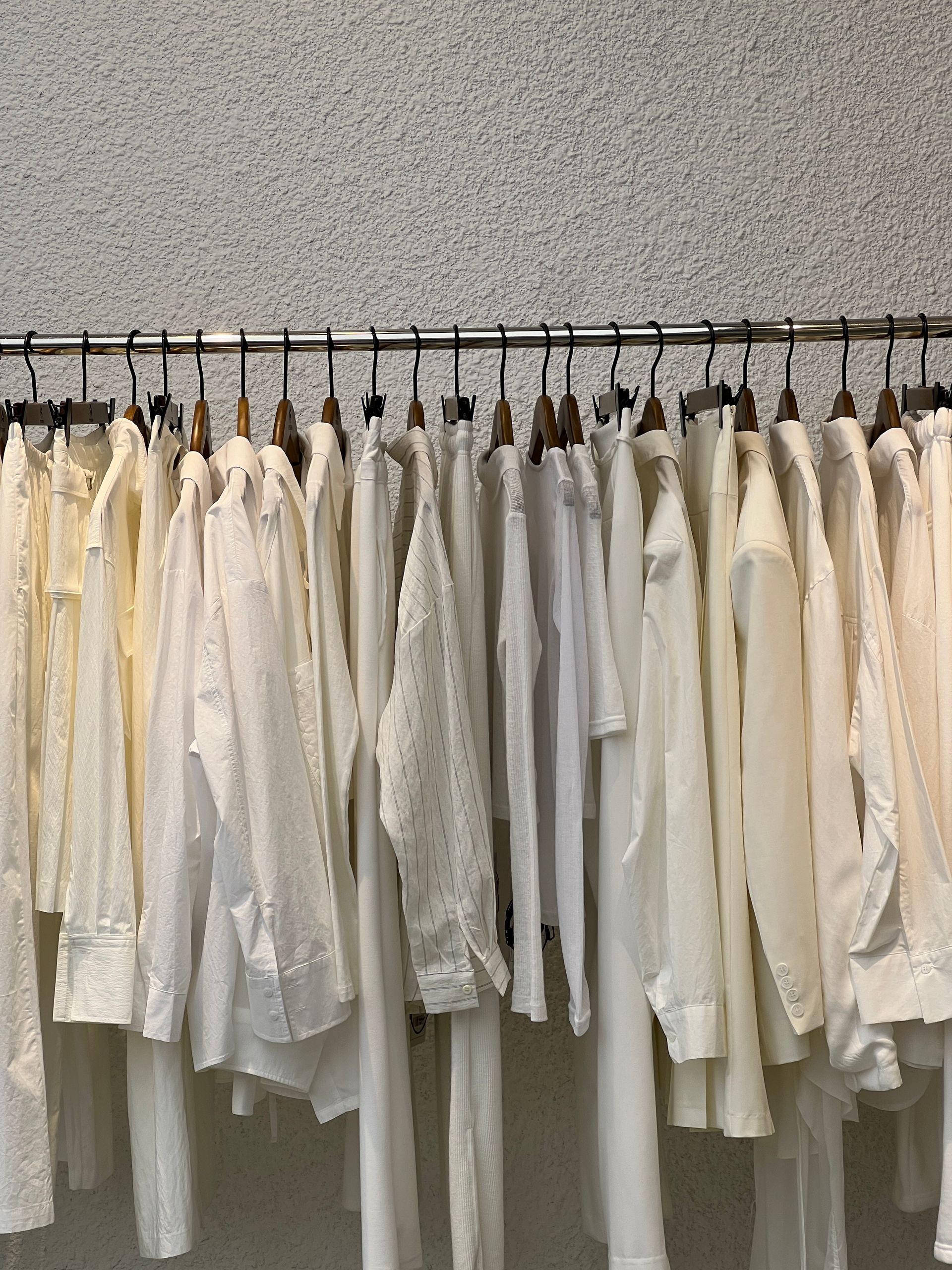 Un perchero con camisas blancas y color crema cuelga contra una pared gris texturizada.
