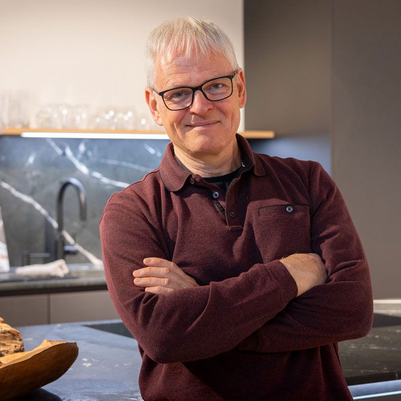 Mann mit Brille steht mit verschränkten Armen in einer Küche und trägt einen weinroten Pullover.