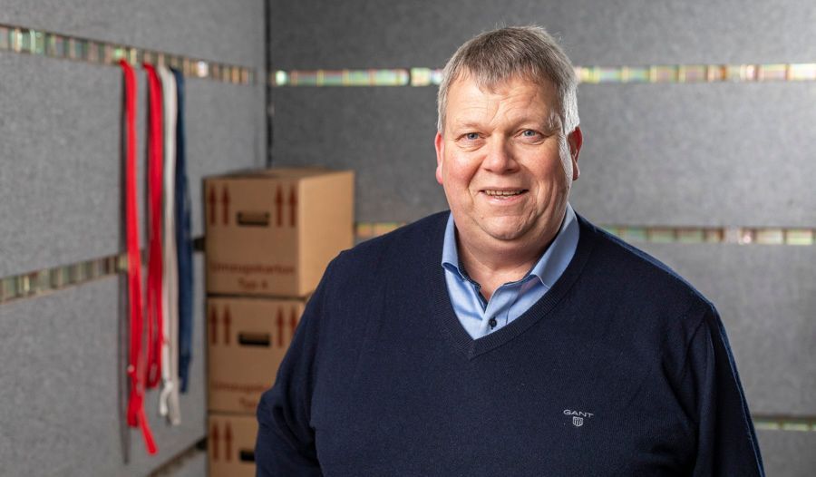 Ein Mann in einem blauen Pullover steht in einem Raum mit Kisten an der Wand.