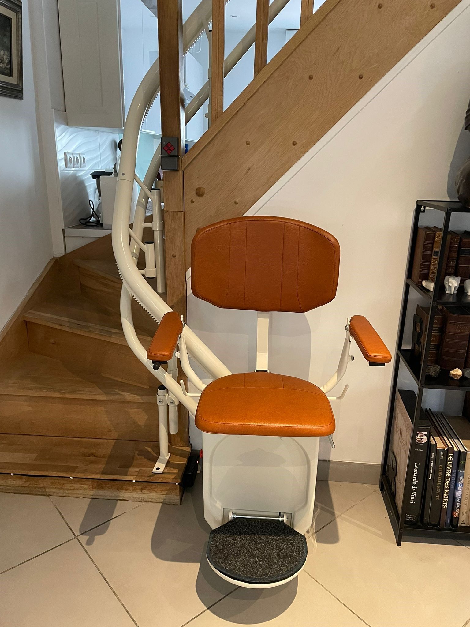 Fauteuil monte-escalier orange sur un escalier avec rampe blanche et repose-pieds repliable dans un intérieur de maison