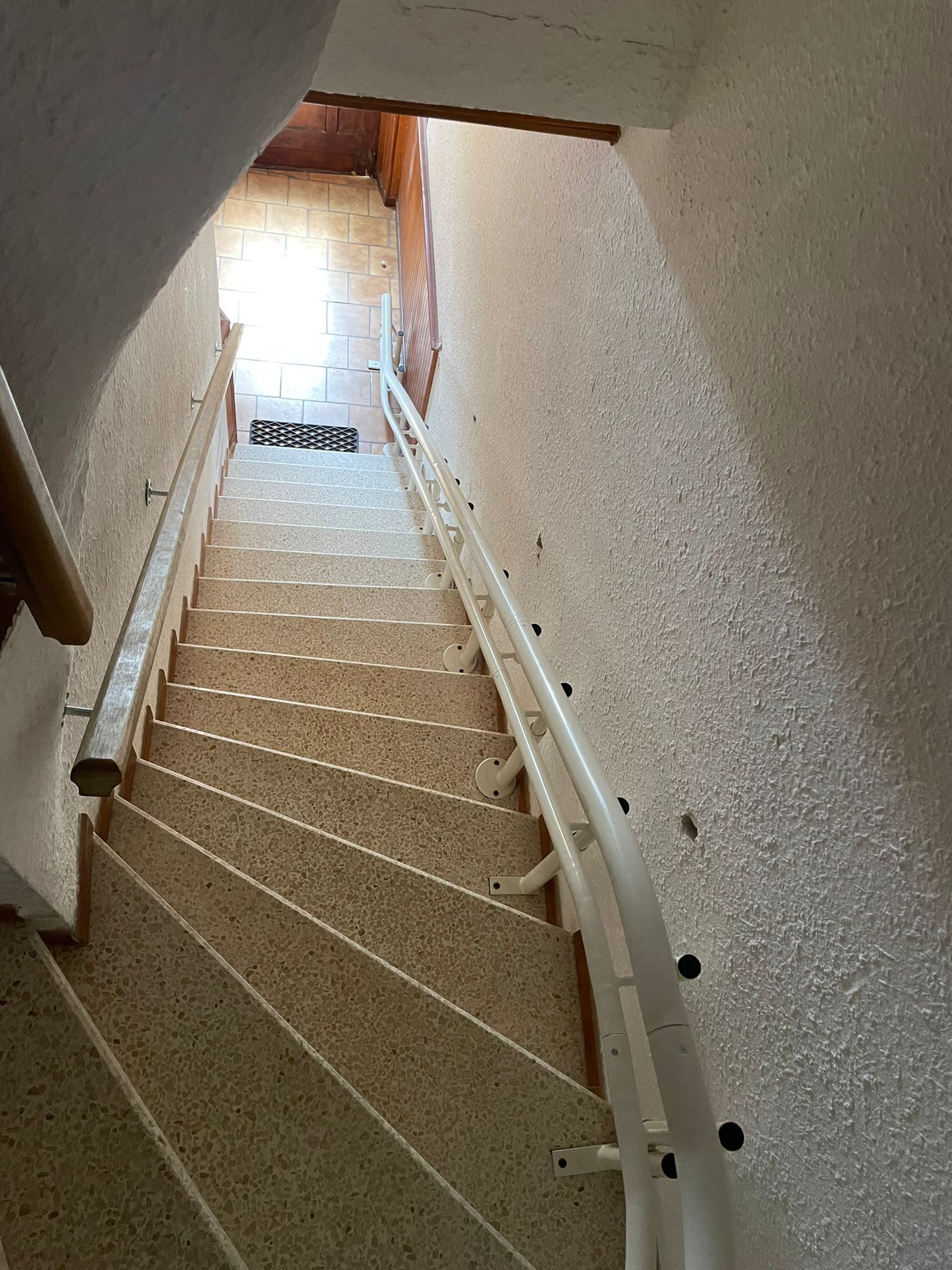 Escalier intérieur raide avec moquette beige, murs blancs et palier lumineux en bas.